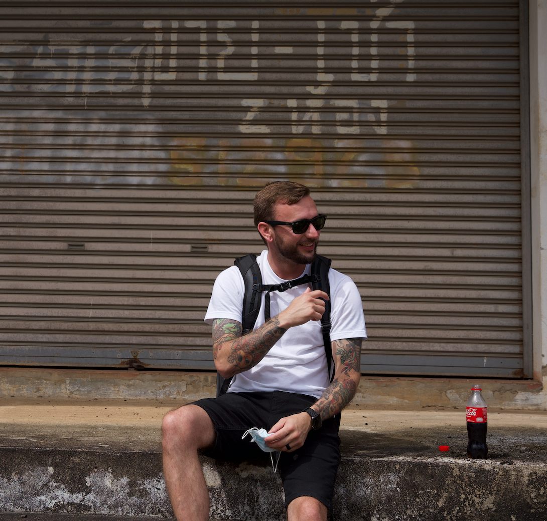Chris sitting on a curb in front of a Thai shop shutter, backpack on, Coke bottle beside him